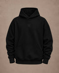 A black pullover hoodie with a small tonal embroidered 'NM' logo on the center chest, displayed against a textured warm brown background.