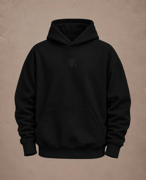 A black pullover hoodie with a small tonal embroidered 'NM' logo on the center chest, displayed against a textured warm brown background.